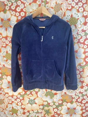 Blå velour hoodie från Juicy Couture - Mjuk och skön blå velour hoodie från Juicy Couture med dragkedja framtill, två fickor och huva. Ikoniskt JC-märke broderat på bröstet och Juicy-text i huvkanten. Perfekt för en chill och trendig look.