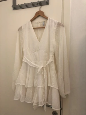 Vit kortklänning med inbyggda shorts från Amisu - Supersöt vit kortklänning från Amisu i storlek 36. Klänningen har v-ringning, långa transparenta ärmar och volanger i flera lager med även inbyggda shorts. Prickigt mönster i tyget och ett knytband i midjan ger extra detaljer. Dragkedja bak för enkel påklädning. På baksidan finns det lite gul fläck men det syns inte så mycket, bara på när håll. Det syns i bilderna också. :) 