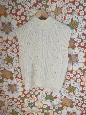 Vit stickad slipover med flätmönster - Stickad slipover i mjuk, krämvit ullmix med höghalsad ribbad krage. Snyggt flätmönster och dekorativa bollar framtill, ribbade kanter och ärmlös design. Perfekt att bära över en skjorta eller t-shirt för en trendig lager-på-lager-look.