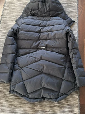 Svart dunjacka från Burton med huva - Snygg svart dunjacka från Burton med huva och dragkedja framtill. Jackan har ett quiltat mönster, en bröstficka med dragkedja och logga på ärmen. Perfekt för kalla vinterdagar och riktigt varm tack vare dunfyllningen. Köpt för 4299kr.