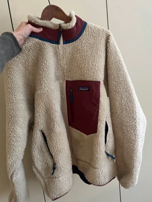 Beige teddyjacka från Patagonia - Mysig beige teddyjacka från Patagonia med vinröda detaljer och blå dragkedjor. Jackan har en stor bröstficka med dragkedja, hög krage och två sidofickor. Perfekt för kalla dagar och riktigt skön att bära. Klassisk och avslappnad passform.