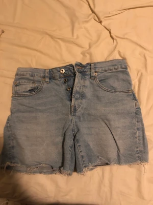 Denim Shorts - Ljusblåa denim shorts. Fyra knappar och gylf. Riktiga fickor fram och bak. Riktiga bältesöglor. Mycket lite stretch.  36 cm långa 84 cm vid midjan/höften 53 cm vid låren
