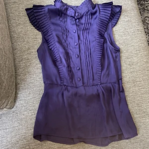 Lila blus - En lila blus med volangliknande detaljer och knappar, ska ha ett skärp eller något liknande men fanns inget när jag köpte det begagnat (Frakt tillkommer)