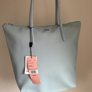 Ny Lacoste axelremsväska  - Helt ny Lacoste väska med läder handtag i pvc vilket är väldigt hållbart. Färger är ljus baby blå och har en när ficka med dragkedja för personliga saker på insidan. Passae jättebra som skolväska. Har haft den i min resväska så lite ”skrynklig” men inga fel. Köpt i Usa för $98.