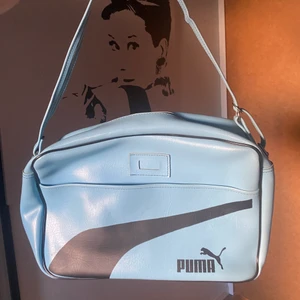 Axelremsväska från puma - Ljusblå axelremsväska från Puma. Knappt använd men köpt från en vintage affär. Väskan är i bra skick, dragkedjan funkar bra och den har inga tydliga defekter annars:) Den är ca 25 cm hög och 45 cm bred