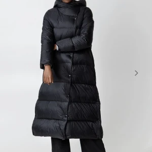 Dunkappa - Lång svart jacka tillverkad i ett vattenavvisande polyestertyg med dunfyllning. Plagget känns lätt och fluffig samtidigt som den är varm. Knäppning framtill och i sidan, samt två sidfickor.