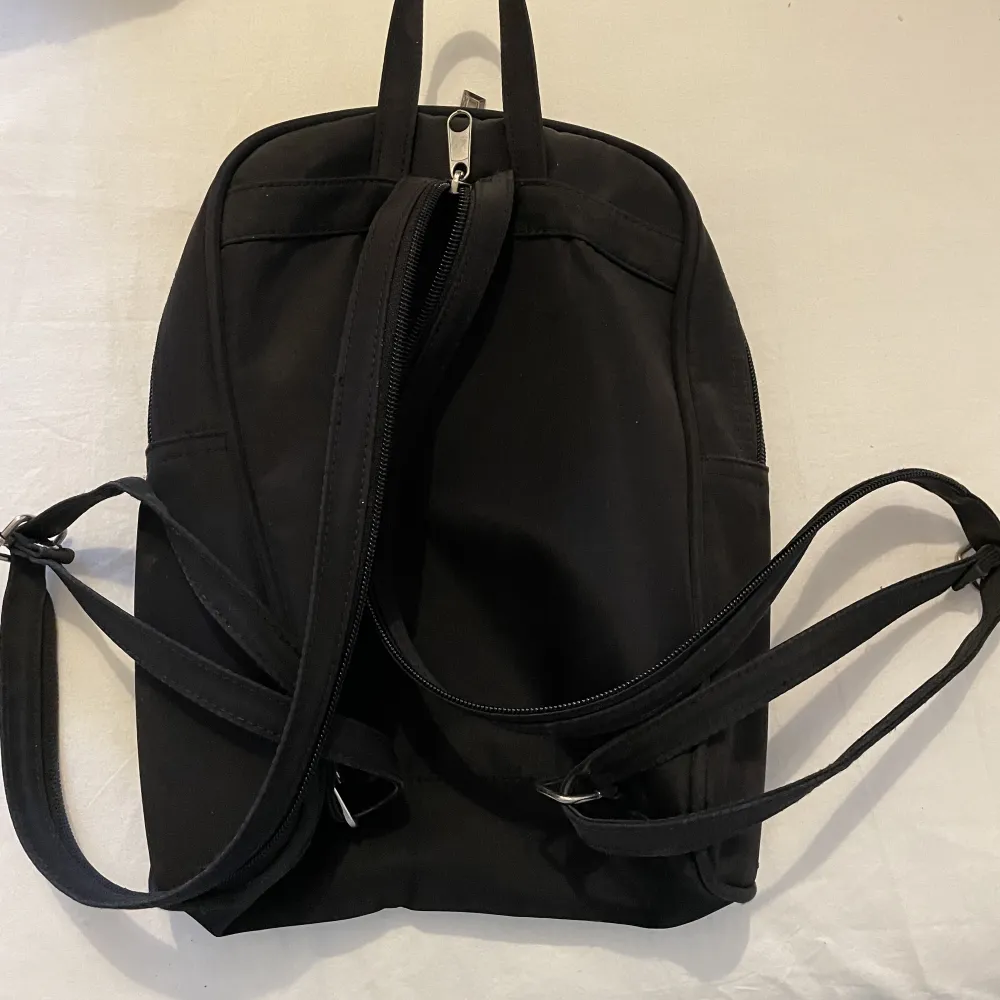 En svart enkel lite ryggsäck med flera fack i och utanför. Med band som går att göra längre eller kortare. 27 cm hög och 25 cm bred.. Väskor.