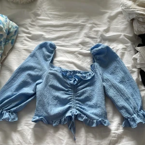 Ljusblå blus  - Supersöt ljusblå blus med 3/4 puffärmar och justerbar dragsko framtill. Volanger både upptill och nedtill. Storlek M (passar S också) 