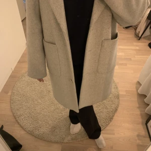 Kappa NA-KD - Jag säljer den här grå/beiga kappan från NA-KD. Superfin modell med två stora fickor. Den är i storlek 40 men passar 42 också. I fint skick men lite nopprig på vissa ställen💕