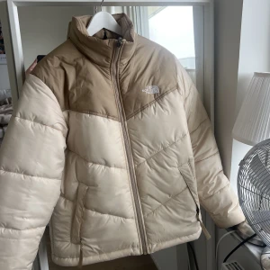 Beige North Face Jacka - Supersnygg dunjacka från the north face! Köpt förra året och enbart använd ett fåtal gånger, vilket också är anledningen till att jag säljer den. Originalpris ligger på 2500kr så vill gärna hålla mig runt 2000kr men jag är villig att ha en dialog om pris.