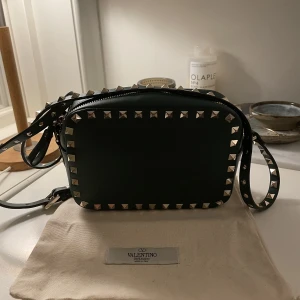 Valentino rockstud camera bag - Säljer min jättefina valentino camera bag i en mörkgrön färg. Älskar den här väskan men har för många väskor. Den har tyvärr tappat tre nitar men man kan gå till valentino och få nya, därav priset. Be om fler bilder på de saknade ställena🫶🏼