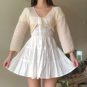 Vintage pointelle-kofta - Söt vintage pointellestickad kofta i ull. Färgen är ljusgul, nästan krämvit och har några ränder i babyblått och ljusrosa nertill. Tunn och mjuk. Fint skick, finns en liten ljus fläck på rosetten som jag inte försökt tvätta bort.