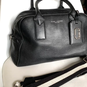 Marc Jacobs väska svart - Svart väska i läder, tillkommer axelrem i svart/vitt. Inköpt i Frankrike ca 2019. Längd 34 cm, bredd ca 14 cm