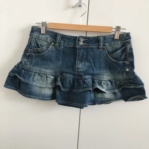 Lågmidjad jeanskjol - Kort lågmidjad jeanskjol med volang nertill, syrrans gamla i nyskick. Stark y2k-stil, perfekt för sommaren! Storleken är 8 vilket verkar motsvara S. Lite stretch i midjan. Mått, midja: 38,5 cm, längd: 28 cm.