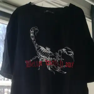 En svart t-shirt med en skorpion som tryck och inbroderat texten 