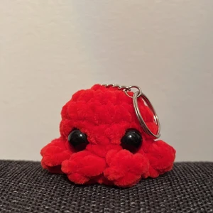 Virkad minibläckfisk-nyckelring - Egengjord, virkad minibläckfisk med nyckelring, i nyskick. Garnet är 100% polyester. Den är ca 6 cm lång och 5 cm hög. Den kostar 39kr totalt med frakt!