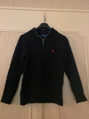  Polo Ralph Lauren tröja  - Kabelstickad svart tröja från Polo Ralph Lauren med halv dragkedja och röd broderad logga på bröstet. Tröjan har ribbade muddar och hög krage, perfekt för kyliga dagar. Klassisk och stilren design som funkar till det mesta.