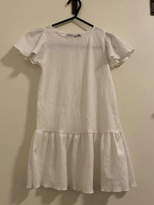 Vit volangklänning med puffärm - Söt vit klänning med puffärmar och volang nedtill. Tillverkad i ett lätt, strukturerat tyg som ger en avslappnad men ändå stilren look. Perfekt för dig som gillar enkel men trendig stil.