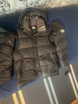 Svart dunväst från Burberry - Snygg svart dunväst från Burberry med avtagbar huva och dragkedjeförsedda fickor. Klassisk quiltad design och ikoniskt rutigt foder i huvan. Perfekt för lager-på-lager och streetwear-stil. Materialmix med bomull och polyamid.