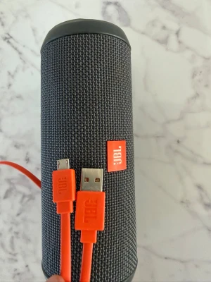 JBL bärbar högtalare svart - Snygg bärbar högtalare från JBL i svart tygmaterial med cylinderformad design. Kommer med orange USB-laddkabel och har en praktisk rem för att ta med överallt. Perfekt för att spela musik trådlöst via Bluetooth.