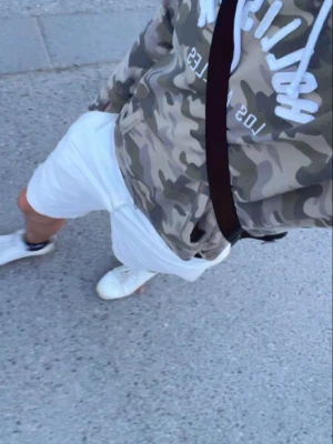 Camo hoodie med tryck från Hollister - Säljer en camouflagemönstrad hoodie från Hollister med vit text 'Los Angeles' på bröstet. Hoodien har huva med vita snören och en stor magficka framtill. Perfekt för dig som gillar streetwear och vill sticka ut lite extra.