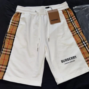 Vita shorts med rutiga sidor - Snygga vita shorts med klassiska beige, röda och svarta rutiga paneler längs sidorna. Shorts med snörning i midjan och logga tryckt på ena benet. Perfekta för sommaren och riktigt stilrena. Passar M med. 