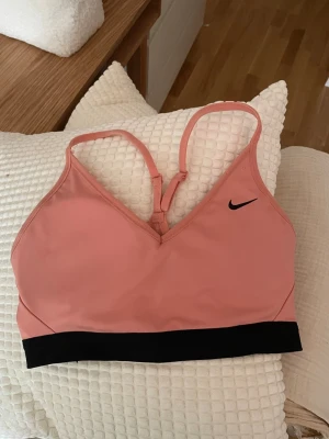 Ljust korallfärgad sport-bh från Nike - Säljer en sport-bh från Nike i storlek M med svarta detaljer och justerbara axelband. Bh:n har en V-ringning och bred resår under bysten för extra stöd. 50 kr!