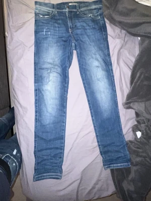 Blå slim fit jeans med slitningar - Säljer ett par blå slim fit jeans med snygga slitningar och tvättade detaljer. Jeansen har klassisk femficksmodell, raka ben och kontrastsömmar på bakfickorna. Perfekta för en avslappnad och trendig look.