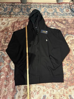 Svart hoodie från Polo Ralph Lauren - Snygg svart hoodie från Polo Ralph Lauren med dragkedja framtill och klassisk huva med snören. Ikoniska loggan broderad på bröstet. Tillverkad i mjukt bomullsmaterial och har ribbade muddar vid ärmar och nederkant. Perfekt för en chill och stilren look.