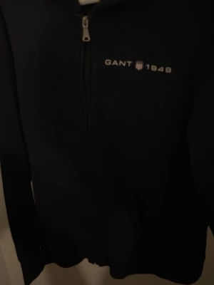Svart hoodie med dragkedja från GANT - Snygg svart hoodie från GANT med halv dragkedja framtill och broderad logga 'GANT 1949' på bröstet. Tillverkad i mjukt material och har långa ärmar. Perfekt för en chill och stilren look.