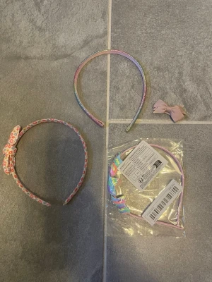 Sött paket med hårband och rosett - Paket med tre hårband och en lös rosett. Ett hårband är regnbågsfärgat med metallicglans, ett har blommigt tyg med rosett och ett är holografiskt med rosett, fortfarande i förpackning. Den lilla rosetten är ljusrosa och kan fästas i håret.