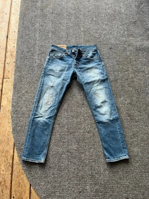 Blå slitna jeans med raka ben - Säljer ett par blå jeans med raka ben och distressed detaljer på framsidan. Jeansen har klassisk femficksmodell och knappgylf. Materialet är denim i bomull och de har en cool, sliten look som passar till många stilar. Passar om man är mellan 163-170 och väger runt 55-63kg