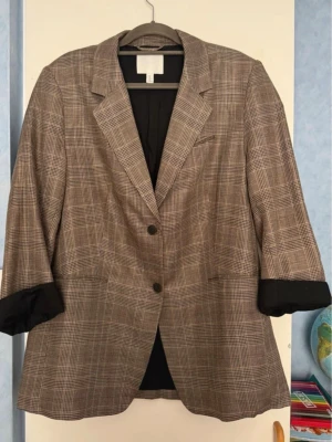 Brunrutig oversized kavaj - Snygg brunrutig kavaj med två knappar framtill och klassisk krage. Kavajen har uppvikta ärmar med svart foder och en avslappnad, oversized passform. Perfekt att styla med jeans eller kjol för en trendig look.