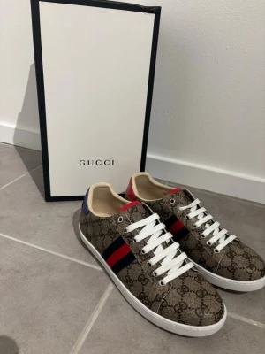 Gucci sneakers med monogram och ränder - Snygga sneakers från Gucci med klassiskt beige-brunt monogrammönster, vita snören och röd-blå rand på sidan. Skorna har vit sula och detaljer i blått och rött på hälen. Perfekta för dig som vill sticka ut med exklusiv stil.