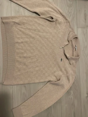 Beige stickad tröja från Lacoste - Säljer en beige stickad tröja från Lacoste med diskret rutmönster och dragkedja vid kragen. Tröjan har lång ärm, ribbade muddar och den klassiska gröna krokodilloggan på bröstet. Perfekt för dig som gillar stilrena och bekväma plagg.