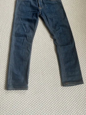 Nudie Hank Rey - Snygga mörkblå jeans med raka ben och klassiska kontrastsömmar. Jeansen har fem fickor och detaljerade bakfickor med broderad söm. Tillverkade i kraftig denim som ger en tidlös look.