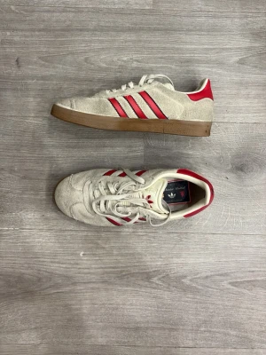 Adidas Hamburg sneakers beige/röda - Snygga Adidas Hamburg sneakers i beige mocka med röda detaljer och klassiska tre ränder på sidorna. Skorna har en brun gummisula och vit insida. Perfekta för dig som gillar retrostil och streetwear.