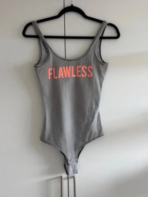 Randig body med texten FLAWLESS - Snygg body med smala svarta och vita ränder samt breda axelband. Framtill står det 'FLAWLESS' i stor rosa text. Stretchigt material och klassisk skärning med rundad urringning. Perfekt för dig som vill ha en trendig och bekväm look.