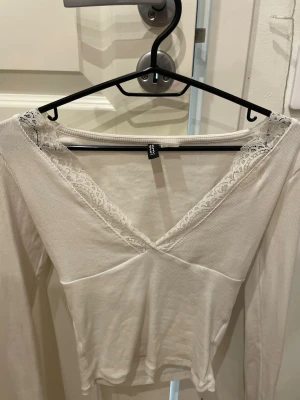 Vit långärmad topp med spets - Säljer en vit långärmad topp från H&M med v-ringning och fina spetsdetaljer runt halsen. Toppen har en omlott-look framtill och är i ett mjukt, ribbat material. Perfekt för dig som gillar en enkel men ändå söt stil.