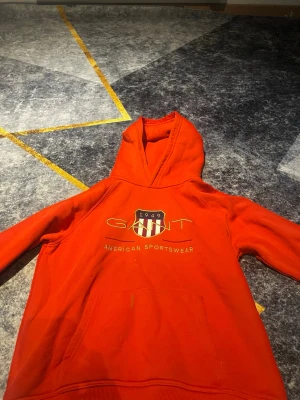 Röd hoodie från GANT med tryck - Snygg röd hoodie från GANT med broderad logga och texten 'American Sportswear' på bröstet. Klassisk huva och stor magficka framtill. Perfekt för en avslappnad och sportig stil. Materialet känns mjukt och bekvämt.