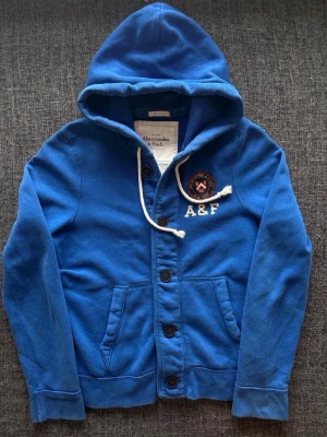 Blå hoodie från Abercrombie & Fitch - Snygg blå hoodie från Abercrombie & Fitch med knappar framtill, vit snörning i huvan och broderad logga på bröstet. Hoodien har två fickor och är perfekt för chill dagar. Klassisk college-vibe och mjukt material.
