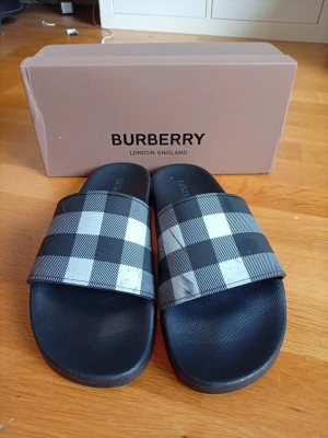 Svarta sandaler från Burberry - Snygga svarta sandaler från Burberry med klassiskt rutigt mönster i vitt och grått på remmen. Tillverkade i syntetmaterial med präglad Burberry-logga på fotbädden. Perfekta för en trendig och avslappnad look.