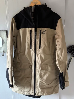 Beige och svart vindjacka Revolution Race - Stilren vindjacka i beige med svarta detaljer från Revolution Race. Jackan har huva, dragkedja framtill, två stora fickor och bröstficka med dragkedja. Justerbara muddar och elastisk midja för skön passform. Perfekt för blåsiga dagar. Helt ny!