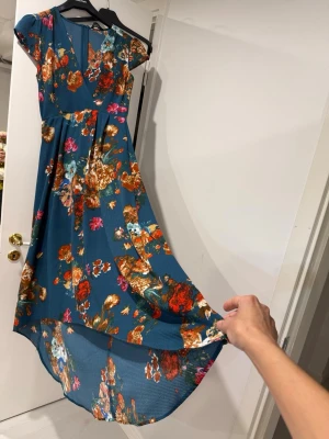 Blommig långklänning i blått - Snygg långklänning i blått med färgglatt blommönster. Klänningen har omlottliv, korta ärmar och lätt v-ringning. Materialet känns som polyester och tyget har en lätt struktur. Perfekt för dig som vill sticka ut med färg och mönster.