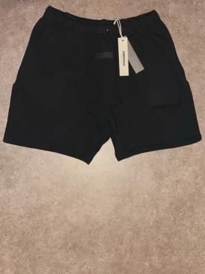 Svarta shorts från Essentials Fear of God - Snygga svarta shorts från Essentials Fear of God med broderad logga på vänster ben. Shortsen har elastisk midja med snörning och är tillverkade i mjuk bomull. Perfekta för en avslappnad och trendig look.