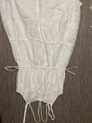 Vit broderad kortklänning med snörning - Söt vit kortklänning med broderade detaljer och snörning i midjan. Klänningen har romantiska volanger och är tillverkad i ett lätt bomullstyg, perfekt för varma dagar. Passar dig som gillar en feminin och avslappnad stil.