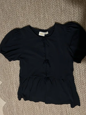 Svart blus med puffärm och knyt - Söt svart blus med puffärmar och två dekorativa knyt framtill. Blusen har en lätt volang nedtill och är tillverkad i ett mjukt tyg som ger en feminin siluett. Perfekt för dig som gillar trendiga detaljer och enkel stil.