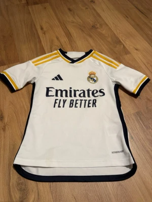Real Madrid matchtröja Adidas - Säljer en vit Real Madrid matchtröja från Adidas med gula och svarta detaljer. Tröjan har klubbmärke och Adidas-logga på bröstet, samt 'Emirates Fly Better' tryckt framtill. Tillverkad i lätt och ventilerande material, perfekt för fotbollsträning eller match.