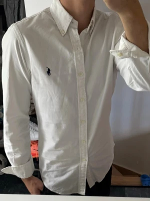 Vit skjorta med knappkrage från Polo Ralph Lauren - Klassisk vit skjorta från Polo Ralph Lauren med lång ärm, knappkrage och ikonisk marinblå logga broderad på bröstet. Tillverkad i bomull för en skön och stilren look. Perfekt till jeans eller chinos för en clean outfit. Lite missfärgad i armhålorna, därav priset. Annars bra skick 