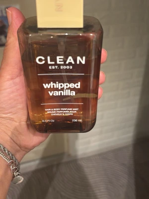 CLEAN Whipped Vanilla Perfume Mist - En hår- och kroppsmist från CLEAN i doften Whipped Vanilla. Flaskan är rektangulär med transparent brun ton och vit text. Innehåller 236 ml och har en enkel, stilren design med beige kork. Perfekt för dig som gillar söta och fräscha dofter.